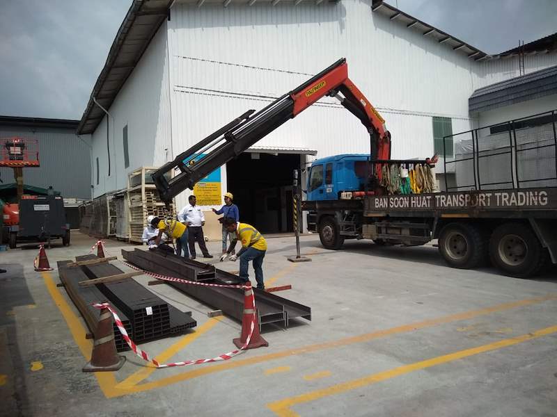 Crane lorry unloading steel sections in the yard, with workers in high-visibility vests and traffic cones - Steel mezzanine platform in a factory (M22) | Wong Lye, BCA General Builder Class 2, Singapore